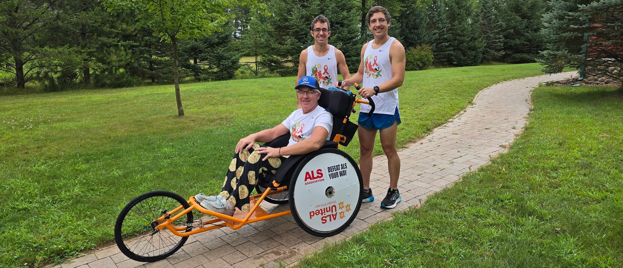 Jeff, Matt, and Mike smiling in their JFB Foundation shirts. Jeff is sitting in an orange racing pushchair wearing his Guinness pajama pants while the boys are standing behind him in running shorts.